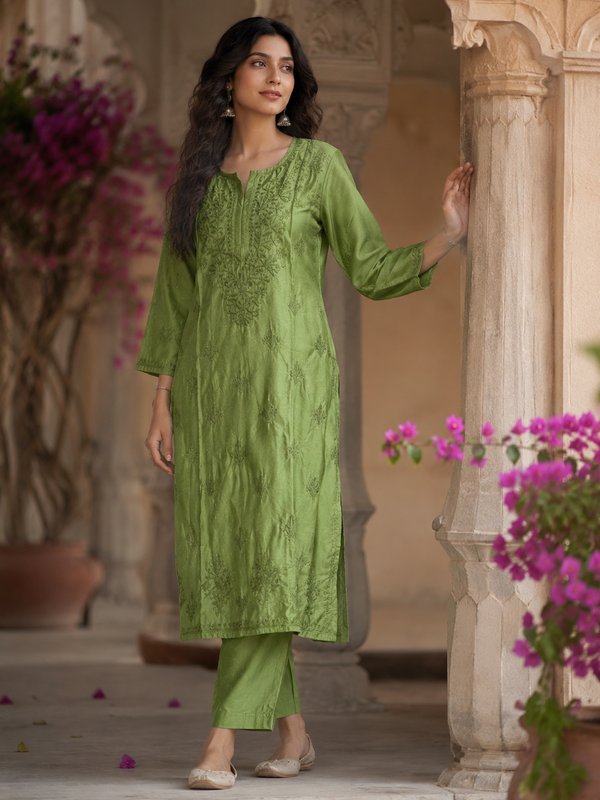 Woman in a green traditional outfit standing in an outdoor setting with flowers and architecture.