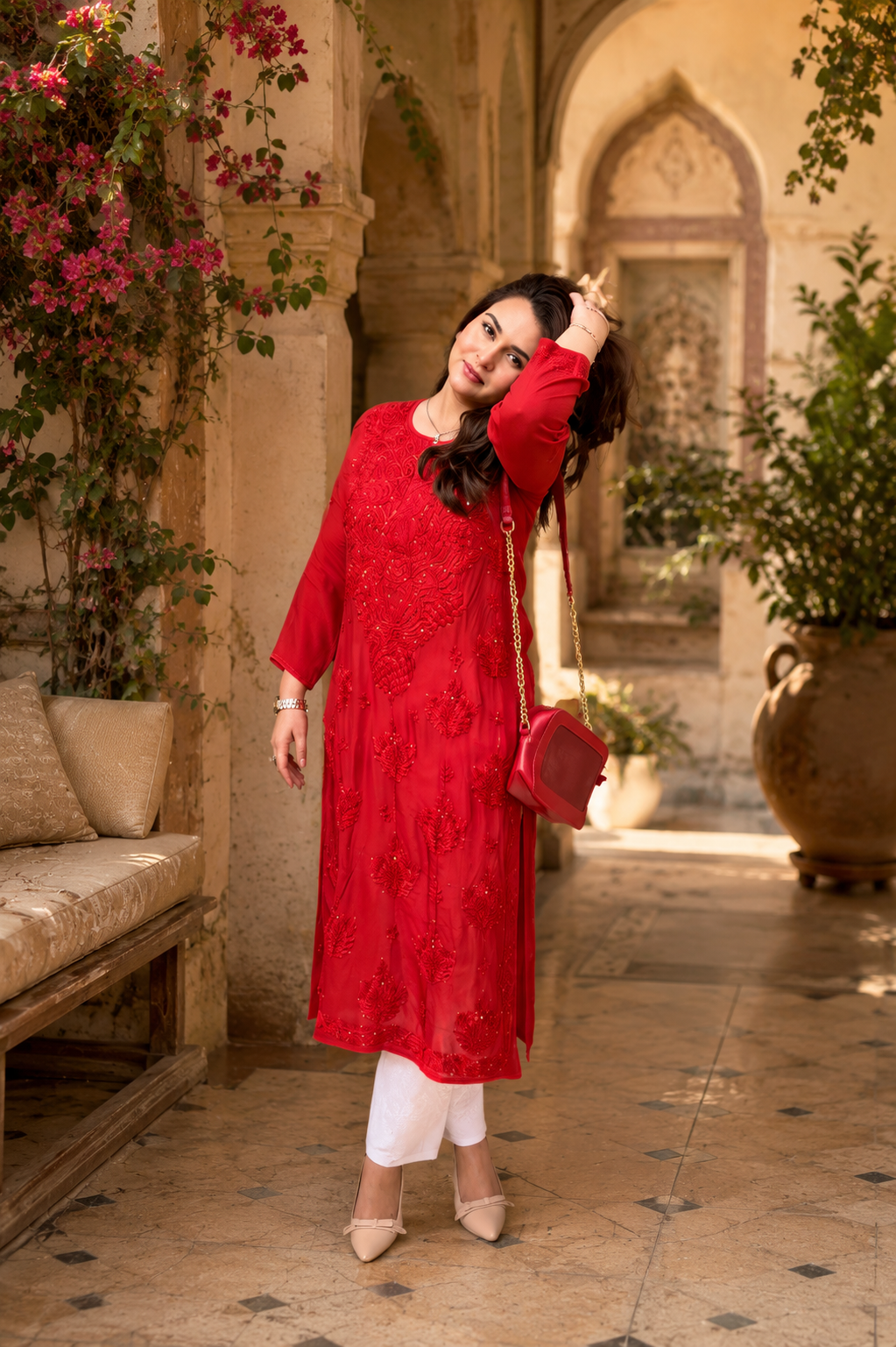Woman in a red traditional outfit standing in an ornate indoor setting with plants and architectural elements.