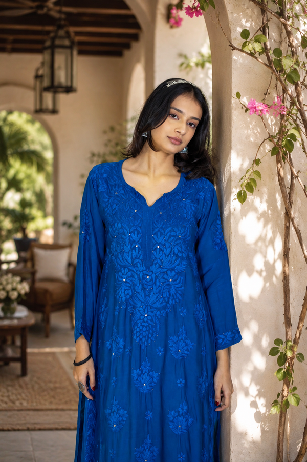 Woman in a blue dress standing in an outdoor setting with plants and furniture.