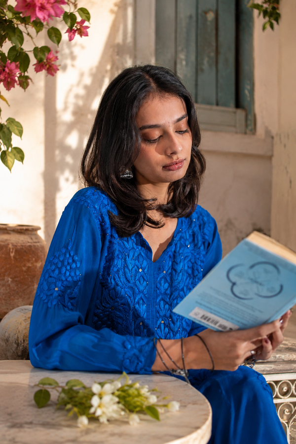Woman in a blue outfit reading a book outdoors with flowers and a building in the background