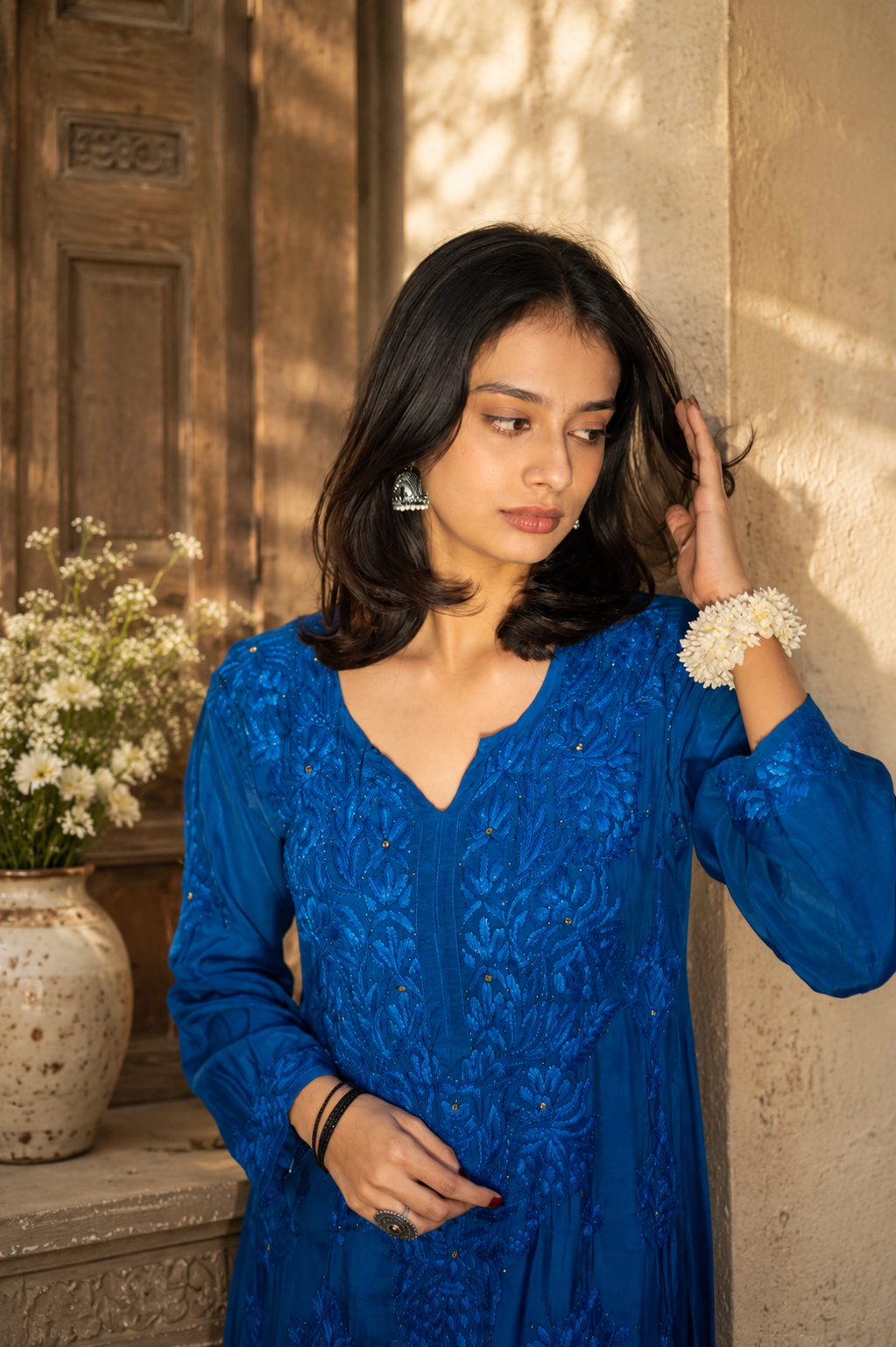 Woman in a blue dress standing against a textured wall with a vase of flowers in the background.