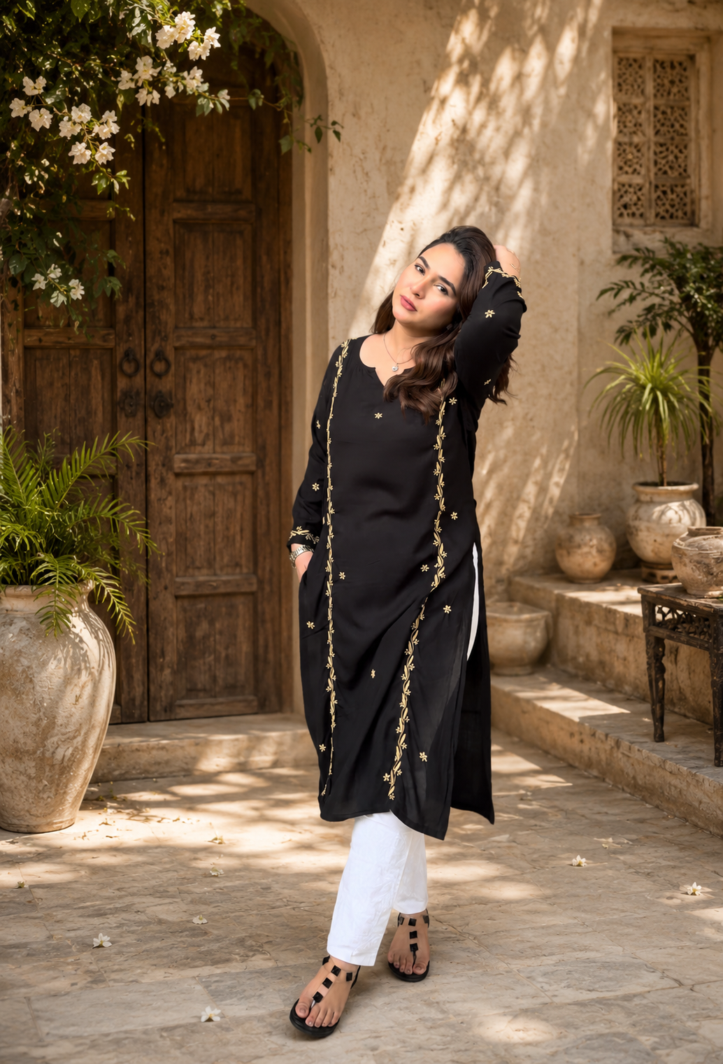 Woman in a black Chikankari traditional outfit standing in a sunlit outdoor setting with plants and a wooden door.