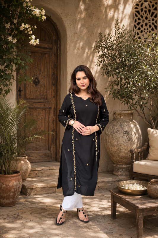 Woman in a black Chikankari traditional outfit standing in an outdoor setting with plants and a wooden door.