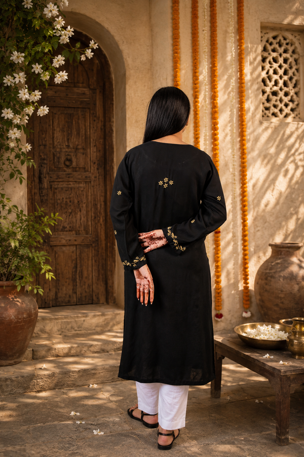 Woman in a black kurta with gold embroidery standing in an outdoor setting with decorative elements.
